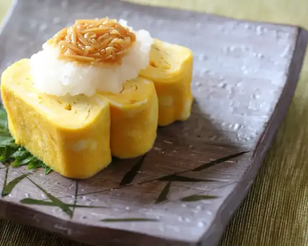 Delikatny omlet tamago, zwinięty w rolki, podany z tartą rzodkwią i grzybami enoki na talerzu z bambusowym wzorem.