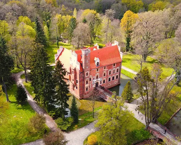 Zamek na wodzie w Wojnowicach otoczony zielenią i drzewami. Czerwona bryła zamku z charakterystycznym dachem wznosi się nad fosą.