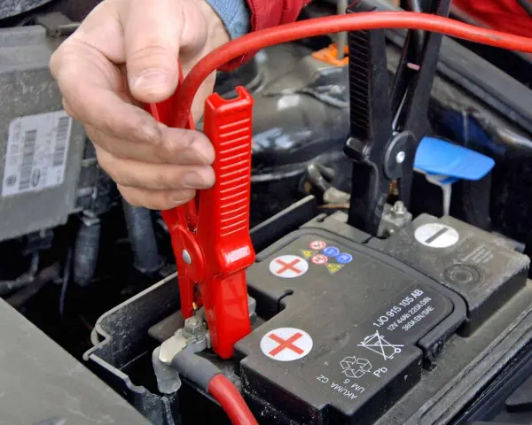 Hand klemmt rote Überbrückungsklemme an die Autobatterie, um Plus und Minus zu verbinden.