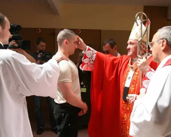 Biskup udziela sakramentu bierzmowania młodemu mężczyźnie. W tle widać osoby z kamerami.