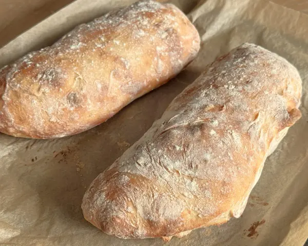 Dwa świeżo upieczone bochenki ciabatty, posypane mąką, leżą na papierze do pieczenia. Idealne do wypróbowania tego ciabatta przepis!