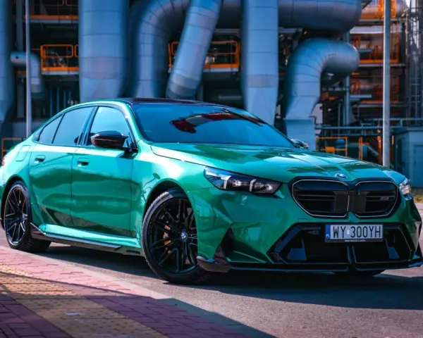 Błyszczące, zielone BMW M5 G90 z czarnymi felgami stoi na tle przemysłowych rur.