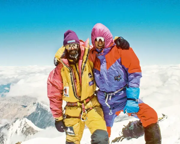 Zwei Bergsteiger auf dem Gipfel, die sich umarmen. Sie tragen bunte Daunenanzüge und Mützen. Die Frage "wie hoch ist der Mount Everest" wird hier beantwortet: 8848 Meter.