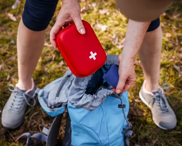 Une personne prépare son sac à dos pour une randonnée, rangeant une trousse à pharmacie enfant rouge avec une croix blanche.
