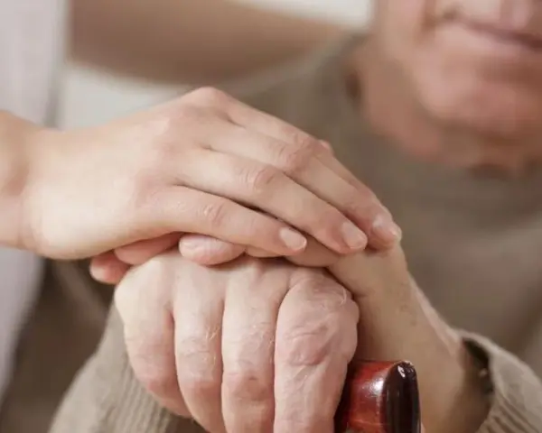 Dłoń opiekunki na dłoni starszego mężczyzny, który trzyma laskę. Wsparcie i troska, jak przy leczeniu np. chorobą Parkinsona, gdzie **Madopar** może pomóc.