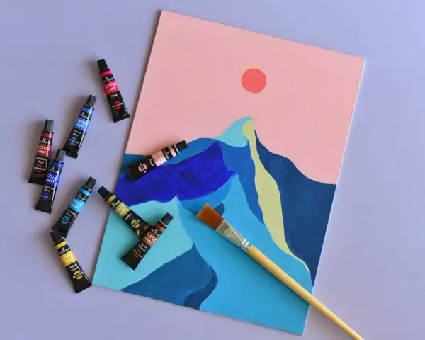 Des tubes de peinture acrylique et un pinceau sont disposés autour d'une toile représentant des montagnes stylisées sous un ciel rose. Une peinture à faire, prête à être créée.
