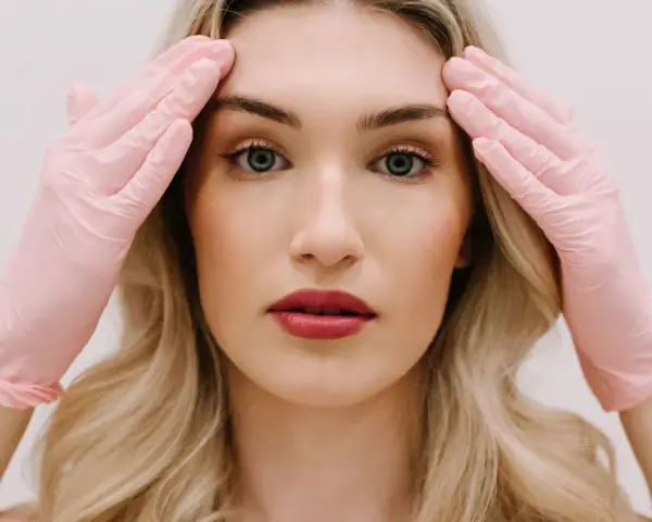 Kobieta z blond włosami w różowych rękawiczkach dotyka skroni. Przygotowanie do zabiegu, jakim jest liposukcja twarzy, by uzyskać idealny kontur.