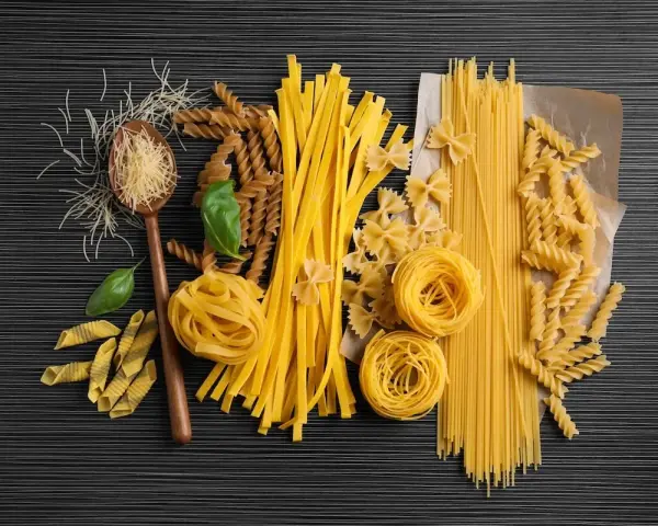 Różnorodne rodzaje makaronu, od cienkich nitek po tagliatelle i fusilli, ułożone na ciemnym tle. Wartości odżywcze makaronu zależą od jego składu.