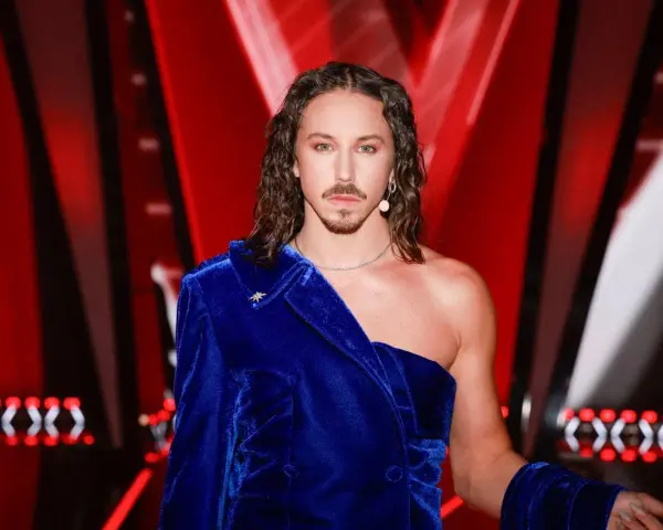 Mężczyzna w niebieskiej, aksamitnej marynarce, z długimi włosami i wąsami, wygląda jakby przygotowywał się do występu na Eurowizji.