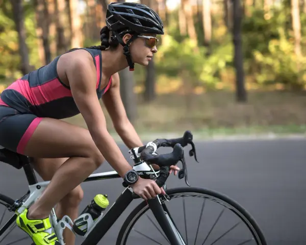 Kobieta w kasku i okularach jedzie na rowerze szosowym przez las. Widać jej nogi w sportowym stroju i zegarek na ręce. Jazda na rowerze - efekty po miesiącu są widoczne w jej sylwetce.