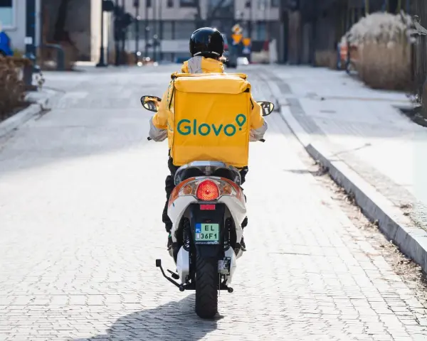 Kurier na skuterze z żółtym plecakiem Glovo jedzie ulicą.