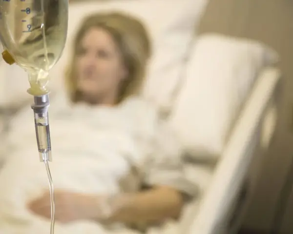 Infusion vorbereiten: Eine Frau liegt im Krankenhausbett, während eine Infusionstropfflasche und ein Tropfschlauch im Vordergrund zu sehen sind.