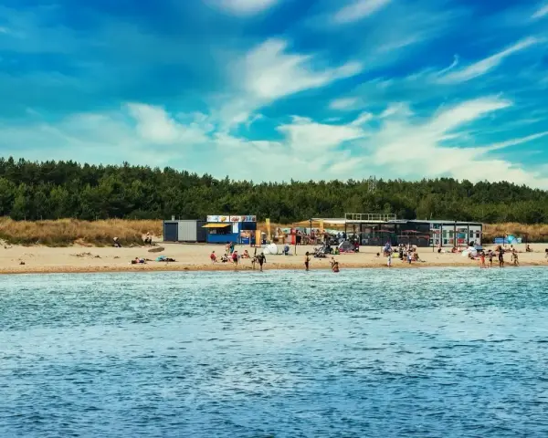 Słoneczny dzień na plaży, ludzie odpoczywają, a w tle widać budynki i las. Idealne miejsce na kemping na Helu.