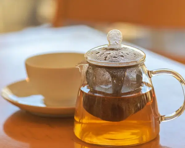 Théière en verre remplie de tisane dorée, avec une tasse en arrière-plan. La question est : combien de temps peut on garder une tisane infusée ?