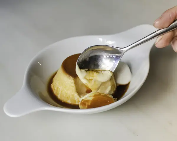 Una cuchara saca un trozo cremoso de flan de cuajada, bañado en caramelo líquido.