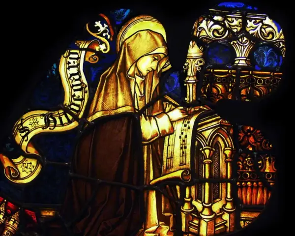Stained glass depicts a cloaked figure, likely the head of a monastery, diligently writing on a scroll.