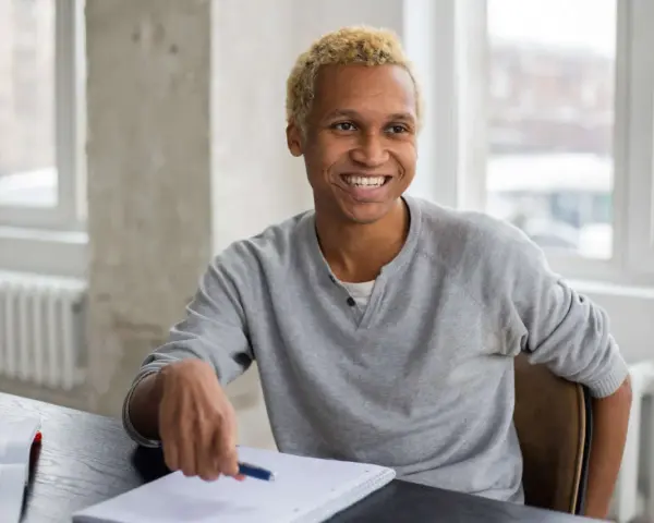 Lächelnder Mann mit blonden Haaren sitzt am Schreibtisch, Notizbuch und Stift bereit für sein Praktikum in Deutschland.