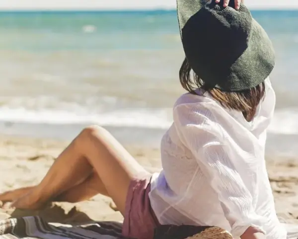 Kobieta w kapeluszu siedzi na plaży, w tle widać spokojne jezioro. Idealne miejsce na ustronny domek nad jeziorem.