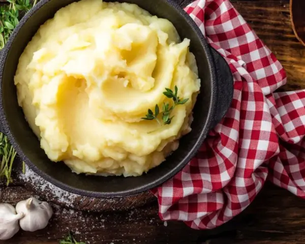 Una ciotola di cremoso purè di patate, guarnito con timo fresco, accanto ad aglio e un tovagliolo a quadri rossi.