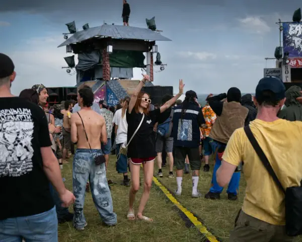 Une foule danse sous la pluie à un festival, une femme au centre lève les bras. On aperçoit une scène et des bannières, peut-être un dj français anime l'événement.
