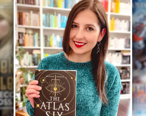 Uśmiechnięta kobieta trzyma książkę "The Atlas Six". W tle widać półki z książkami, w tym inne serie książek fantasy.