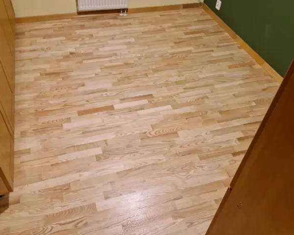 Jasny, odnowiony parkiet po cyklinowaniu, lśniący w świetle. Obok drewniane meble i zielona ściana.