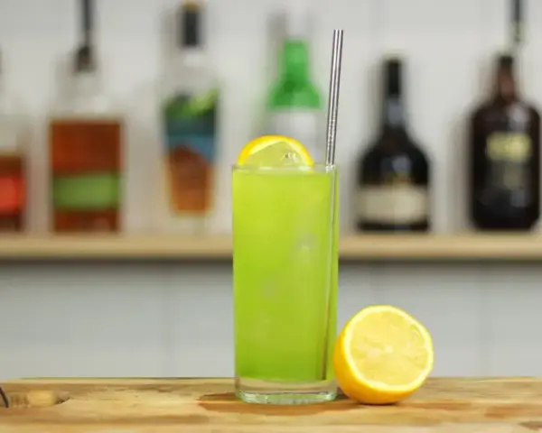 Zielony, orzeźwiający drink Tokyo Iced Tea z plasterkiem cytryny i metalową słomką, na tle półki z butelkami alkoholu.