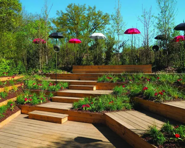 Un jardin en pente moderne avec des terrasses en bois, des parterres de fleurs rouges et des parasols colorés.