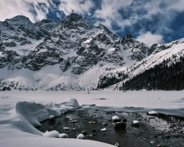 Zamarznięte Morskie Oko, otoczone ośnieżonymi szczytami Tatr. W oddali widać ludzi spacerujących po lodzie, a na pierwszym planie płynie strumyk.