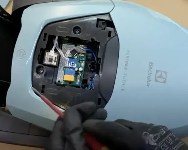 En person med svarta handskar skruvar på ett kretskort inuti en ljusblå Electrolux dammsugare, märkt "PURE D8.2 SILENCE".