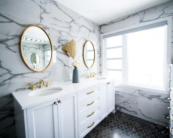 A modern bathroom vanity installation featuring white cabinets, marble walls, gold accents, and dual sinks.