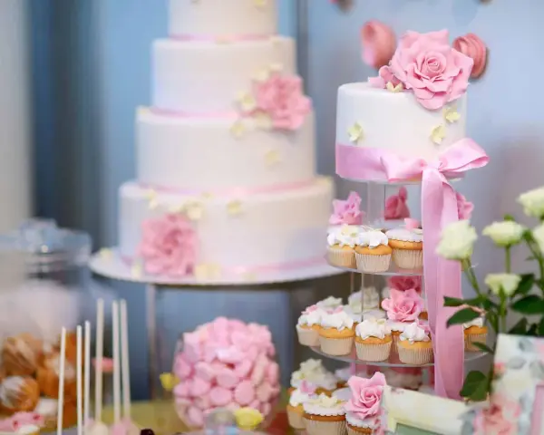 Eleganckie, białe torty weselne i babeczki ozdobione różowymi kwiatami i wstążkami. Idealne na słodki stół, inspiracja jakie ciasta na wesele.