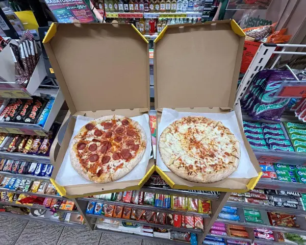 Dwie pizze w pudełkach, jedna z salami, druga serowa. Obok półki ze słodyczami i napojami w sklepie Żabka.