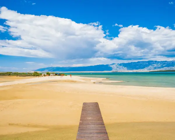 Drewniany pomost prowadzi na piaszczystą plażę Ninskiej laguny. W tle turkusowe morze i góry.