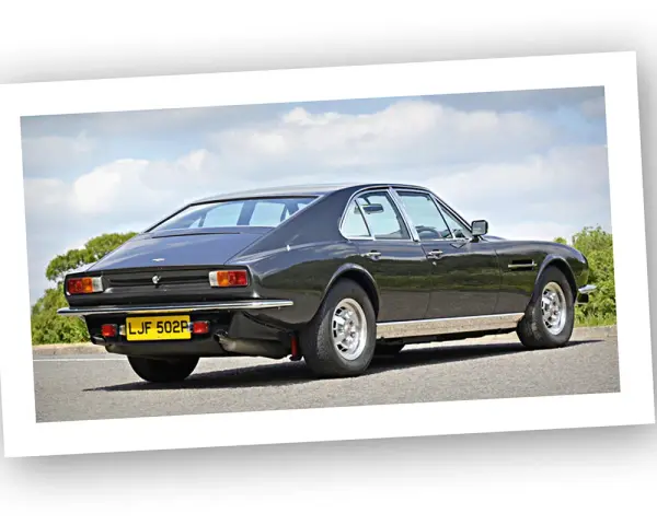 Klasyczny Aston Martin Lagonda w ciemnym kolorze, widziany z tyłu, na tle błękitnego nieba z chmurami.