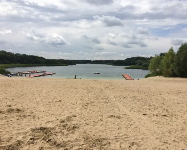 Piaskowa plaża nad jeziorem, z pomostami i łódkami. W oddali las, chmury na niebie. Idealne miejsce na wypoczynek nad wodą.