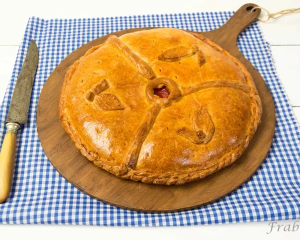 Empanada dorada y decorada sobre tabla de madera, con cuchillo y mantel a cuadros.