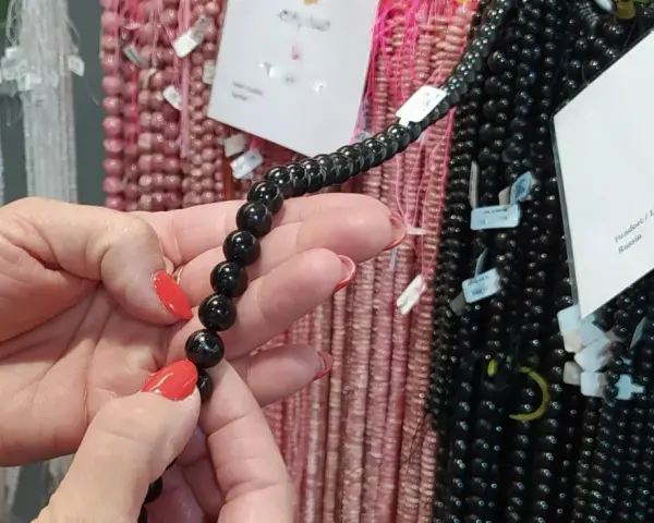 Manos con uñas rojas sostienen un collar de cuentas negras de shungita. En el fondo, se ven hilos de cuentas rosas y blancas.