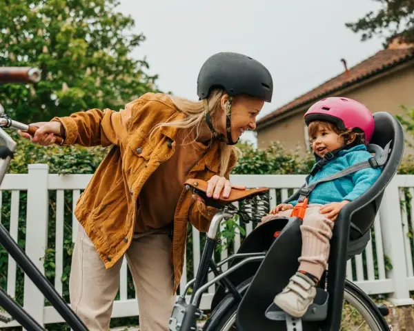 En kvinna med hjälm ler mot ett litet barn i en cykelbarnstol. De är redo att cykla med bebis i det vackra vädret.