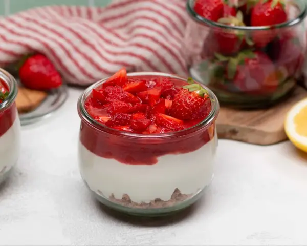 Zwei Gläser mit cremigem Erdbeer-Cheesecake, garniert mit frischen Erdbeeren und Erdbeersauce. Daneben ein Glas voller Erdbeeren und eine Zitronenhälfte.