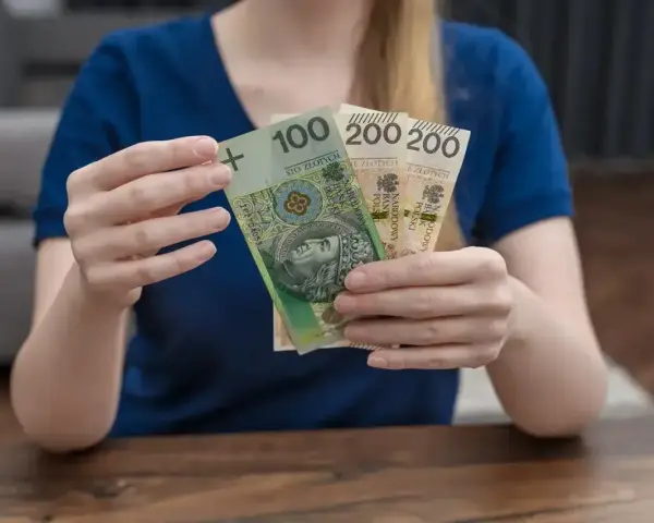 Dłonie trzymają polskie banknoty: 100 zł i dwa razy po 200 zł. To może być część zasiłku rodzicielskiego.