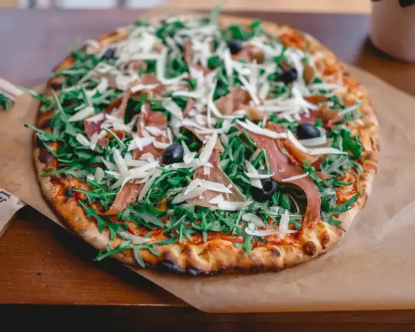 Pyszna pizza prosciutto z rukolą, oliwkami i parmezanem, podana na papierze do pieczenia.