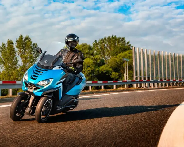 Niebieski Peugeot Metropolis 400 z kierowcą w kasku pędzi po zakręcie drogi, otoczony zielenią i błękitnym niebem.