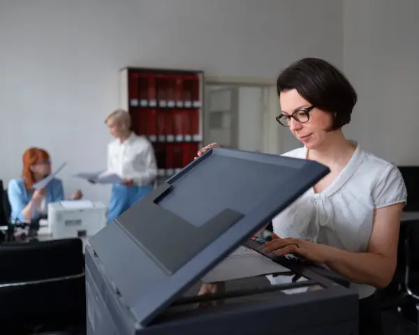 Frau mit Brille bedient einen Drucker, vielleicht um einen Epson Drucker zu installieren. Im Hintergrund arbeiten Kolleginnen.