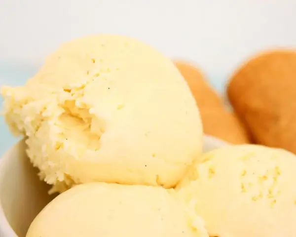 Cremoso helado de vainilla con motas oscuras, servido en un tazón blanco. Al fondo, conos de helado dorados esperan.