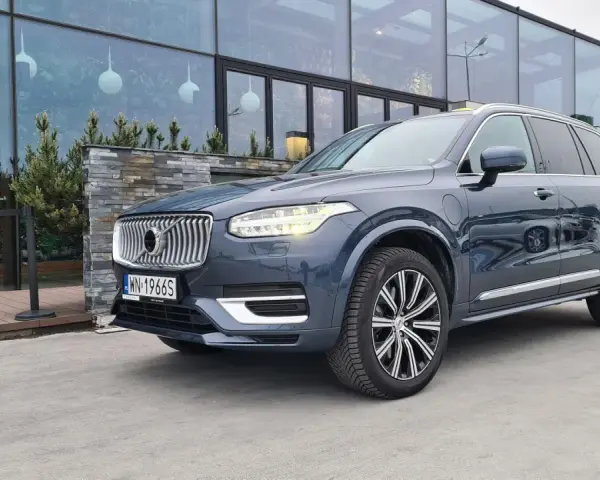 Elegancki, ciemnoszary Volvo XC90 T8 Recharge z polskimi tablicami rejestracyjnymi stoi przed nowoczesnym budynkiem z dużymi oknami.