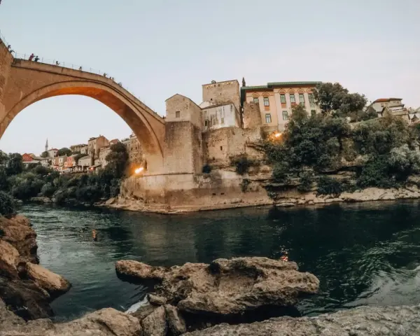 Iconic Stari Most bridge over the Neretva River, a must-see for any Bosnia travel adventure. People watch from the bridge as swimmers enjoy the emerald waters below.