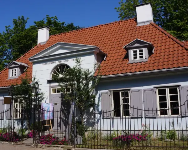 Jasnoniebieski dworek Sierakowskich z czerwoną dachówką, otoczony zielenią i ogrodzeniem.