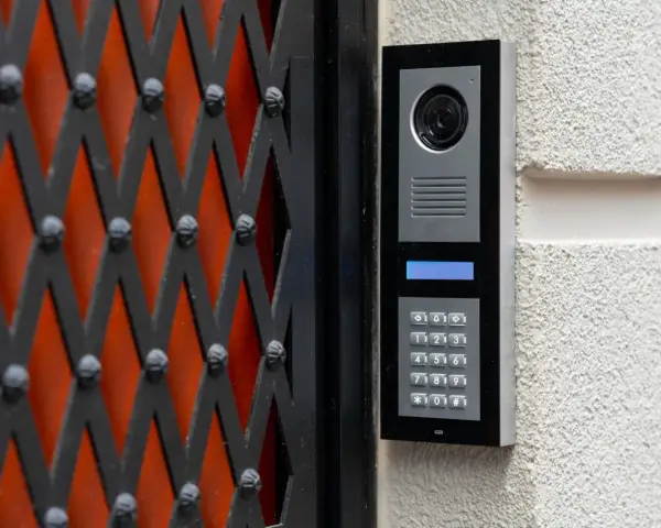 Domofon z kamerą i klawiaturą numeryczną zamontowany na ścianie obok czarnej kraty.
