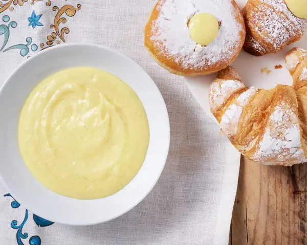Una ciotola di soffice crema pasticcera senza uova accanto a cornetti e bomboloni spolverati di zucchero a velo.